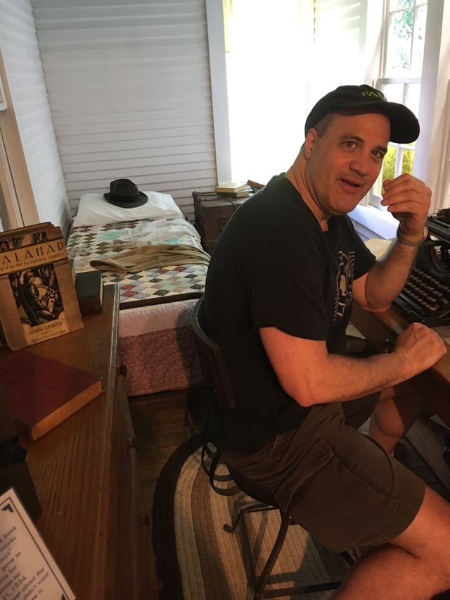 Man sitting at a desk in a room with window blinds, wearing a black cap and shirt, gesturing with his hand.