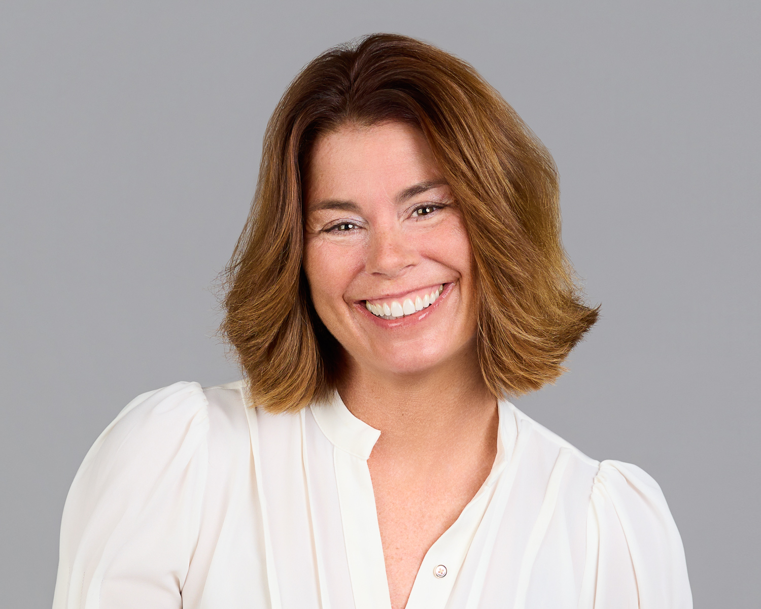 Smiling woman with shoulder-length brown hair wearing a white blouse against a gray background.