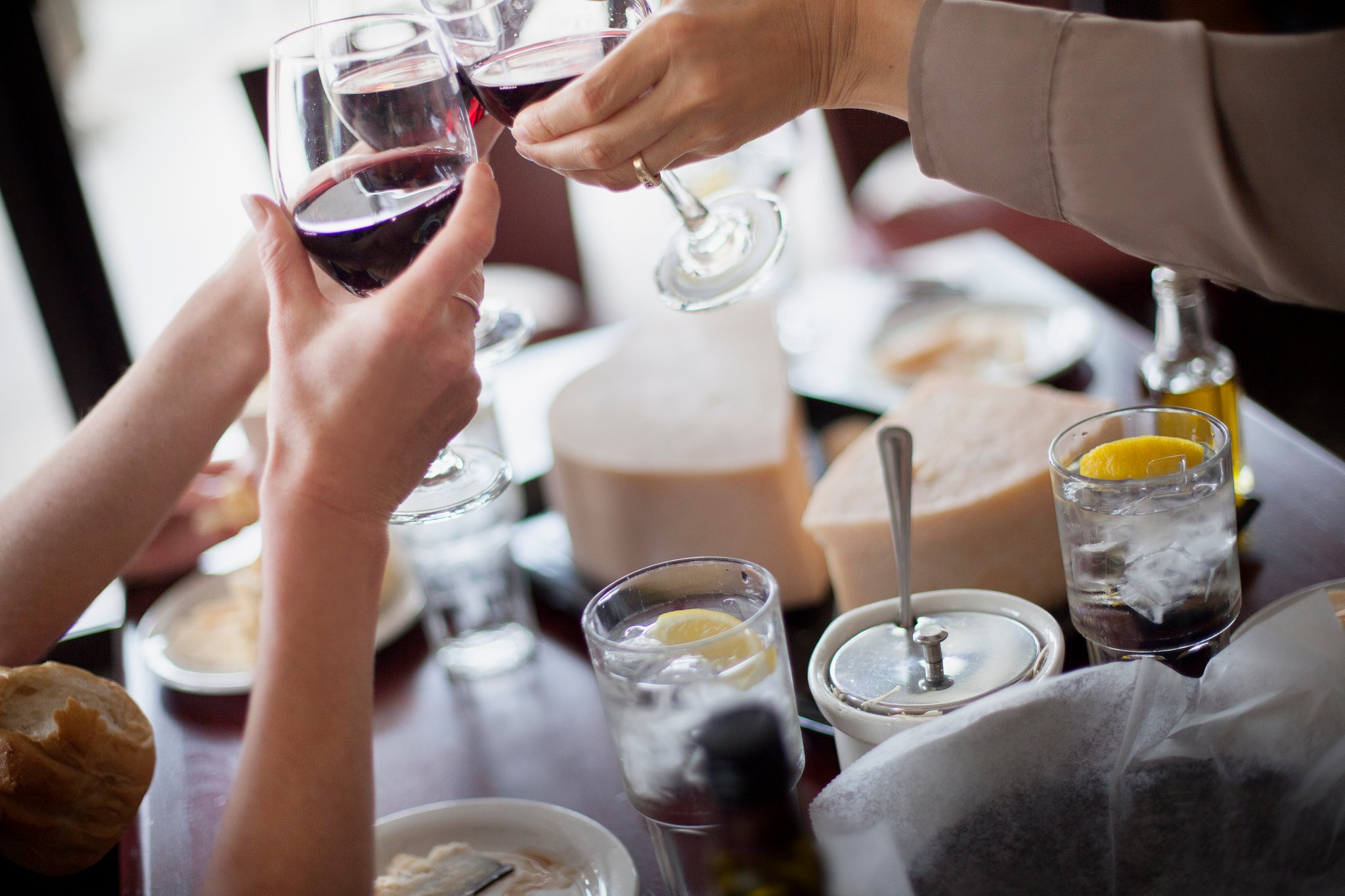 Person holding a glass of red wine, another person pouring wine, table with drinks and food items.