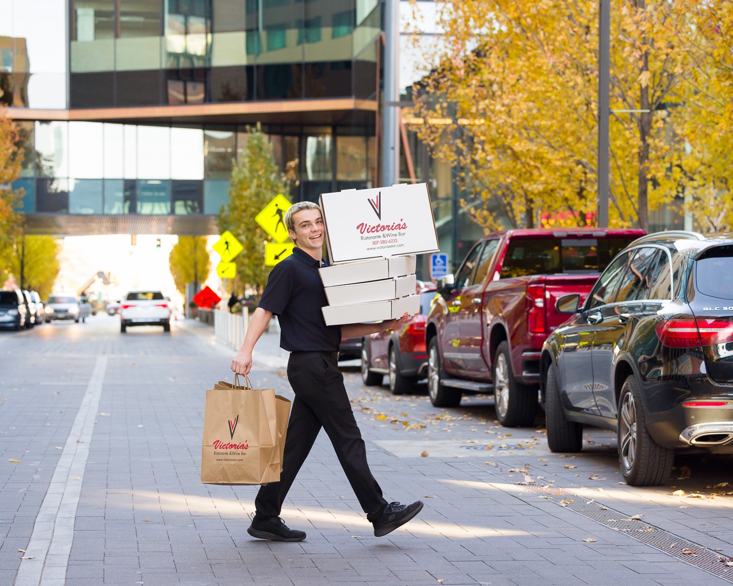 Person walking on sidewalk carrying boxes and a shopping bag, parked cars and trees in background.