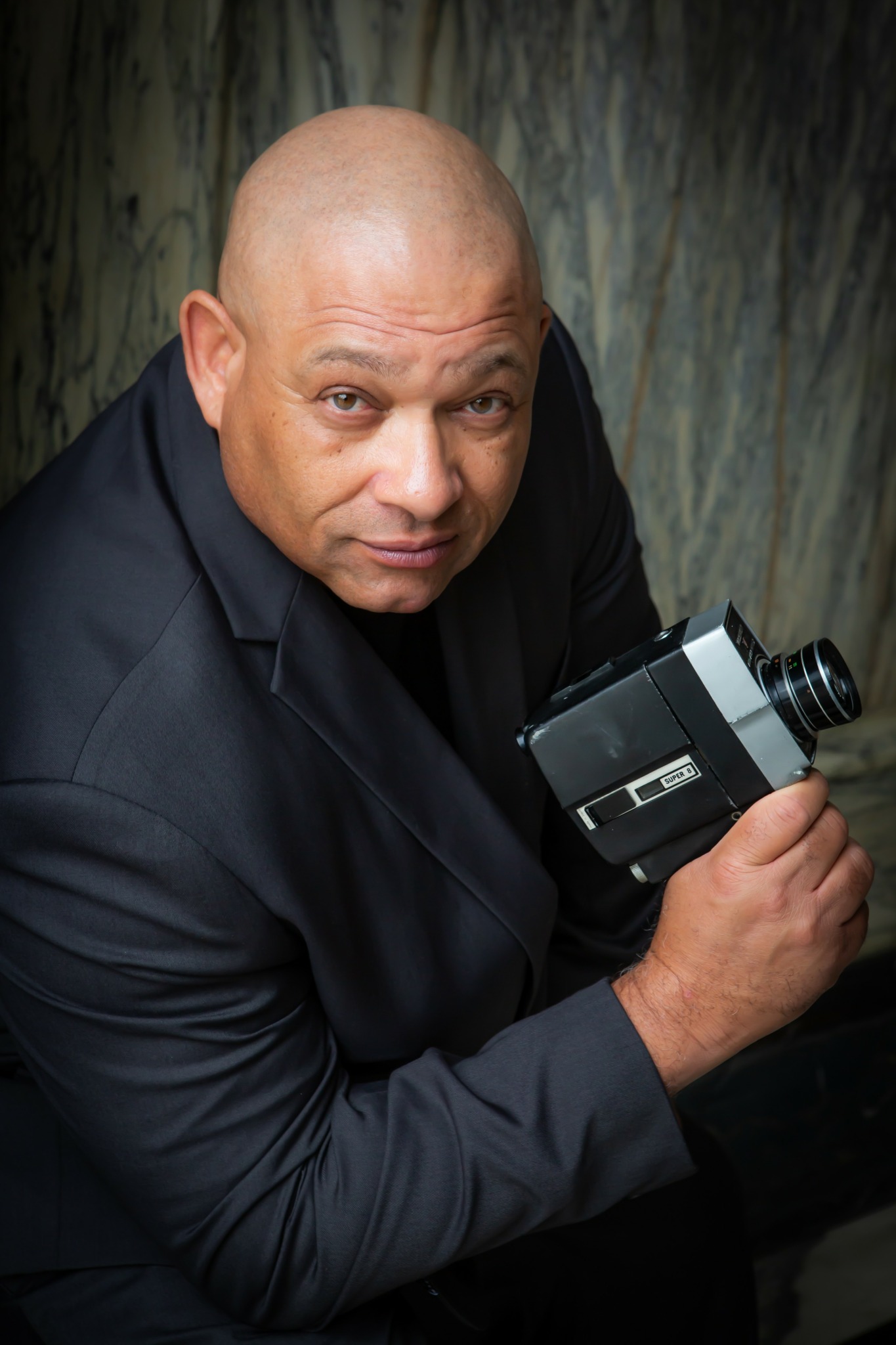 Bald man in dark suit holding a camera, looking up at the camera, against a textured background.