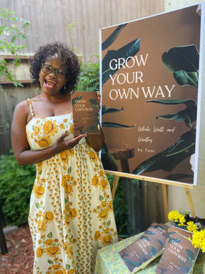 Woman with curly hair and glasses holding a book, smiling, standing outdoors next to a large poster.