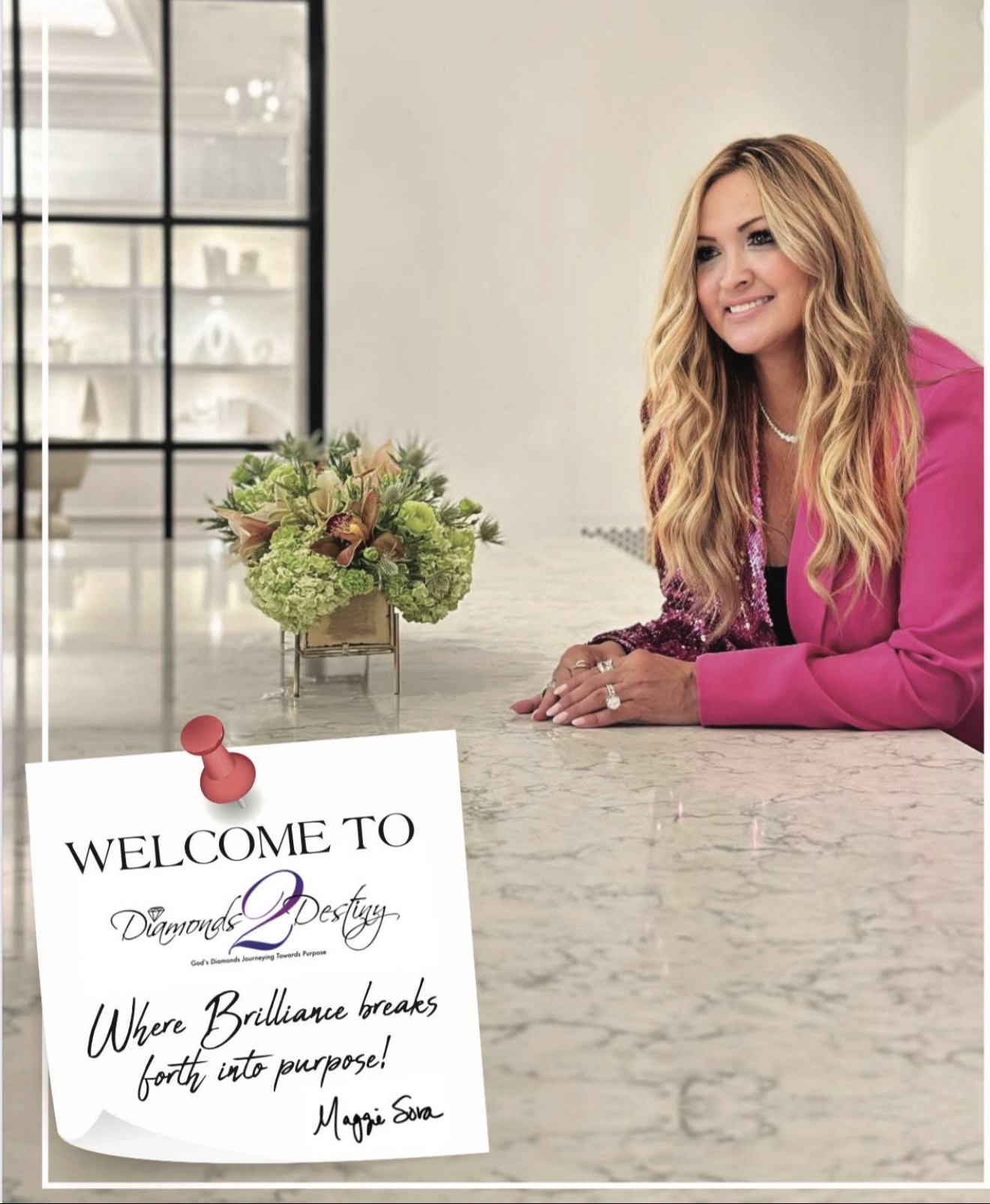 Woman with long blonde hair smiling at a marble counter with a floral arrangement, in a bright room.