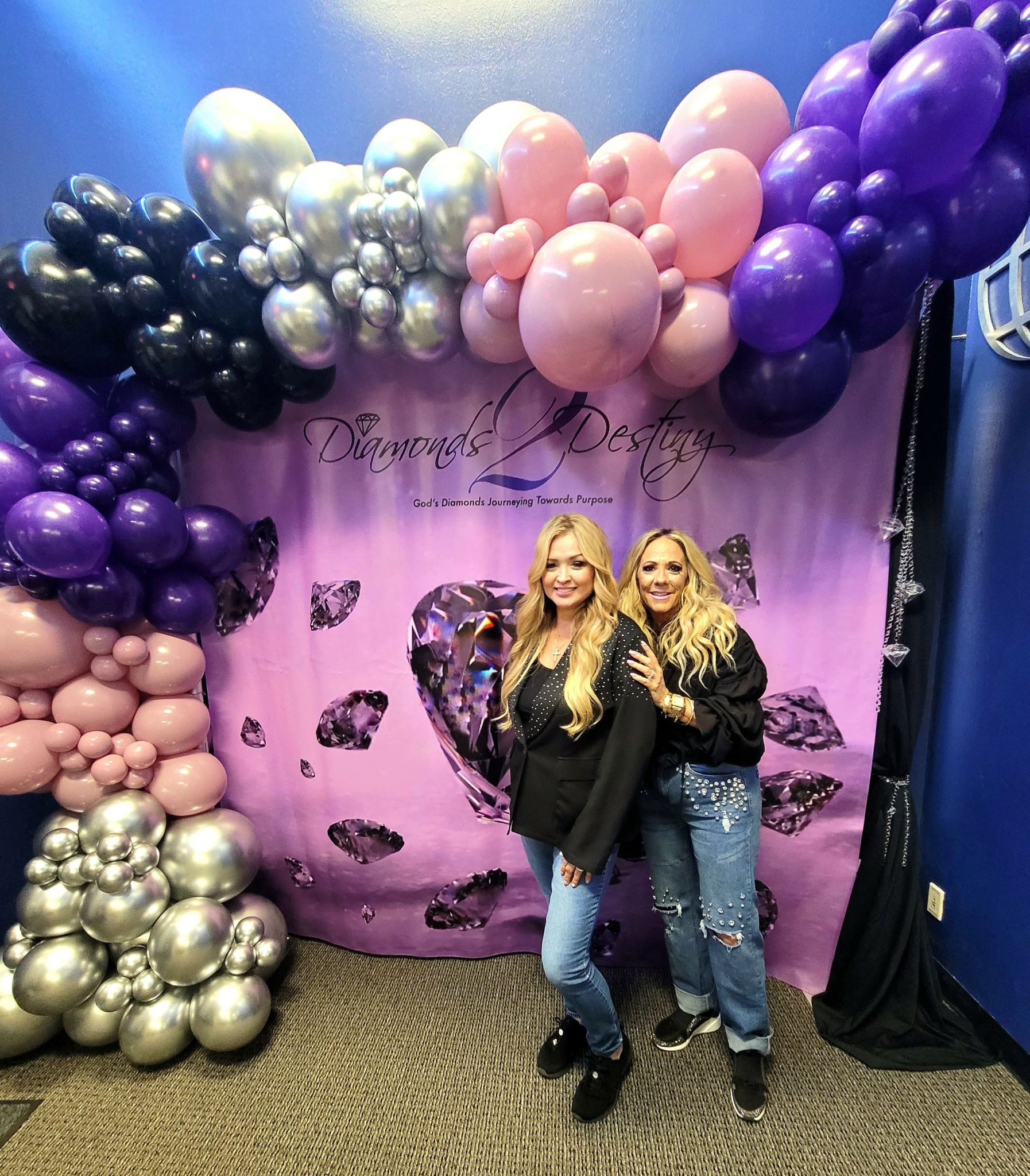 Two women stand in front of a balloon backdrop with pink, purple, black, and silver balloons, and a pink curtain with butterflies.