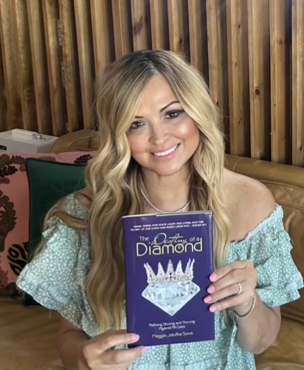 Woman with long wavy blonde hair smiling, holding a blue book titled 'The Diamond' in a cozy indoor setting.