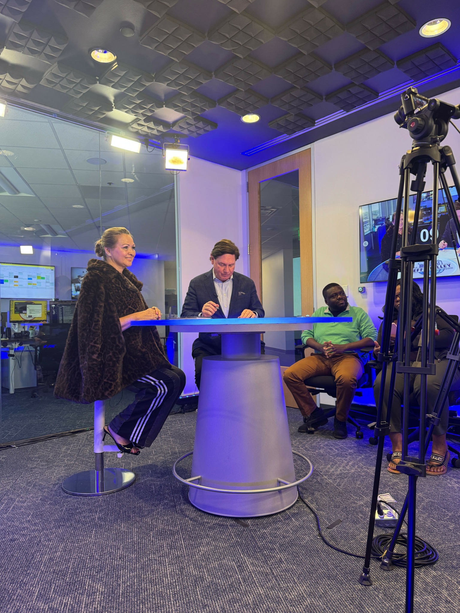 Three people seated around a round table in a studio with camera equipment and a large screen behind them.