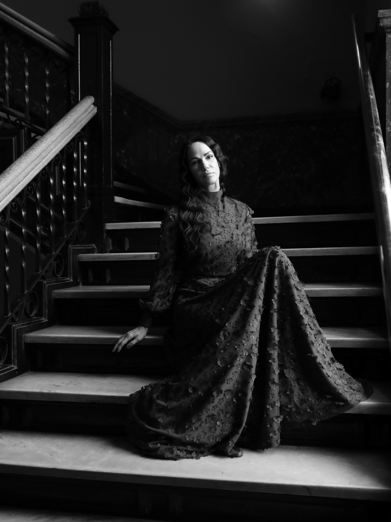 Woman in a long, dark lace dress sitting on stairs in a dimly lit setting.