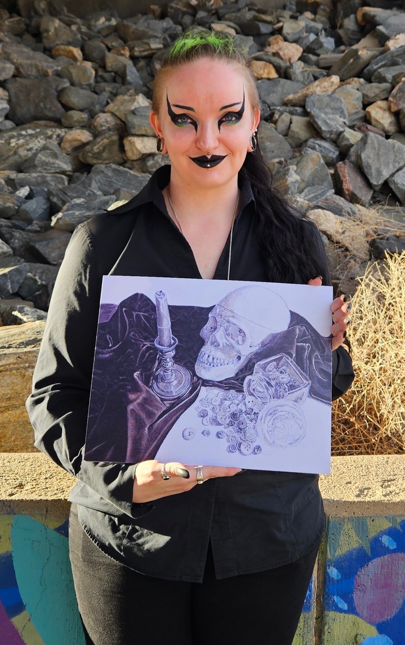 Person with dark makeup and green hair holding a drawing of a skull, bottle, and glass outdoors.