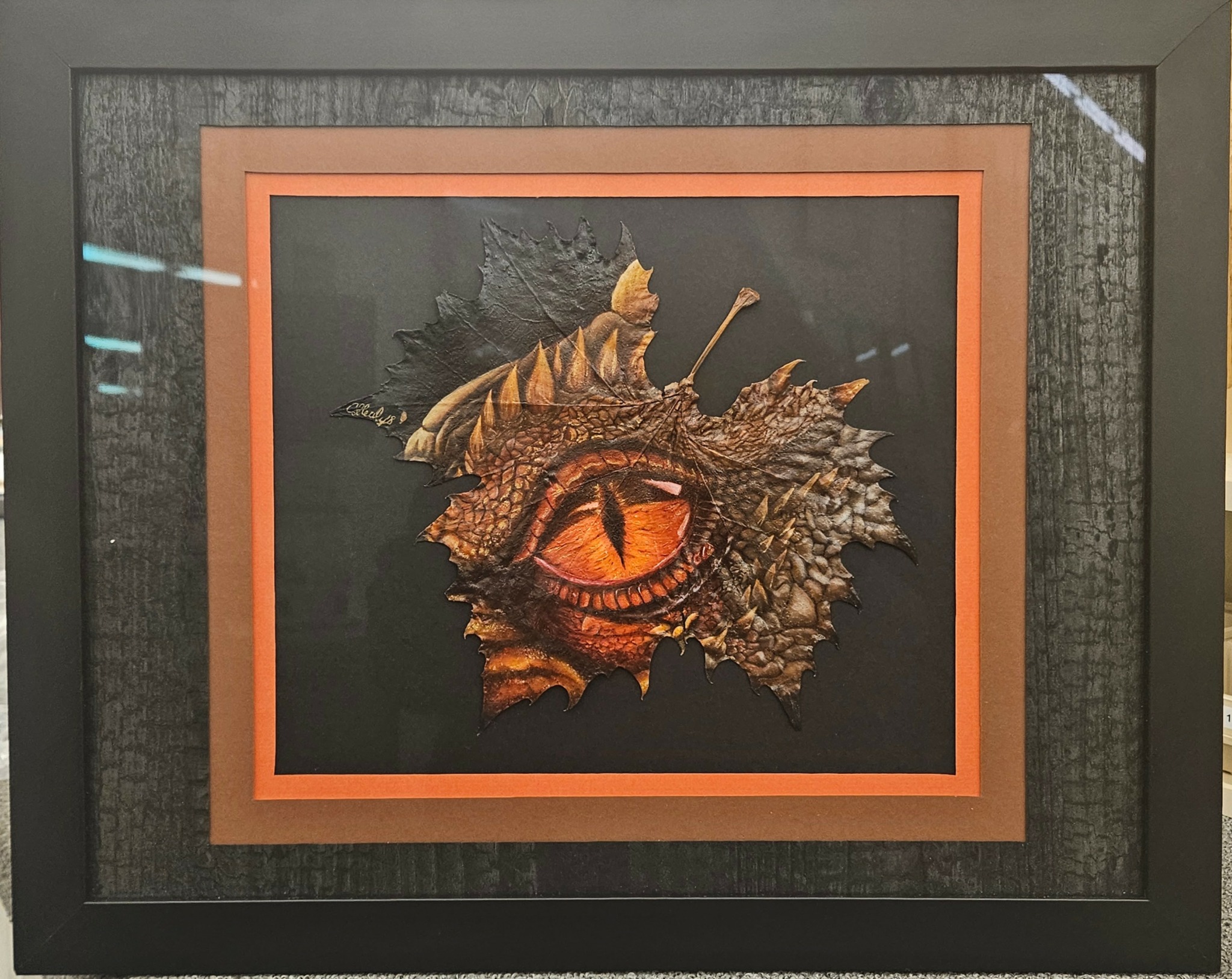 Artwork of a dragon's eye with textured scales and an orange border, framed on a wall.
