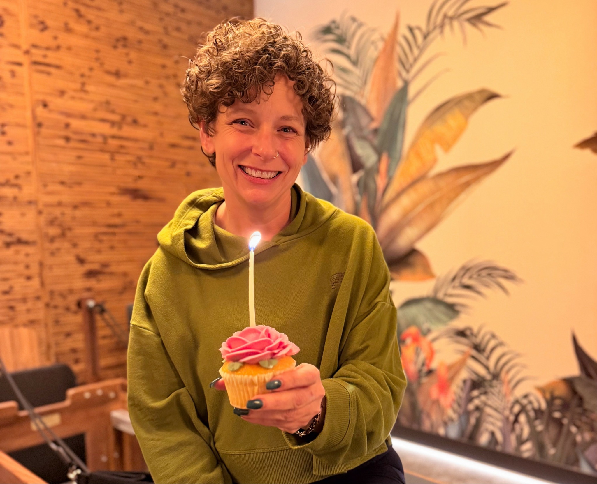 Person with curly hair holding a cupcake with pink frosting and a lit candle, smiling indoors.