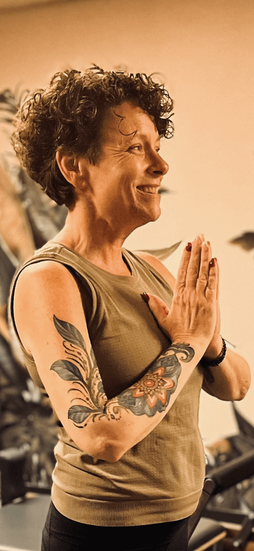 Woman with curly hair smiling, hands pressed together in a prayer position, tattoos on arms, in a gym setting.