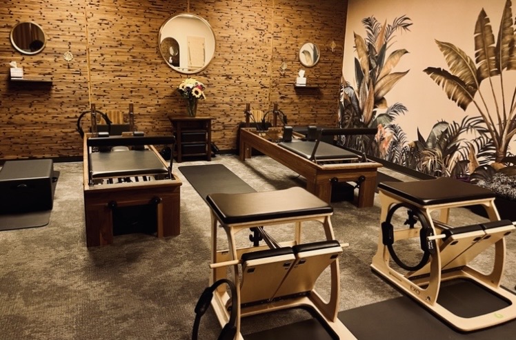 Room with brick wall, mirrors, plants, and Pilates reformer machines arranged in rows.