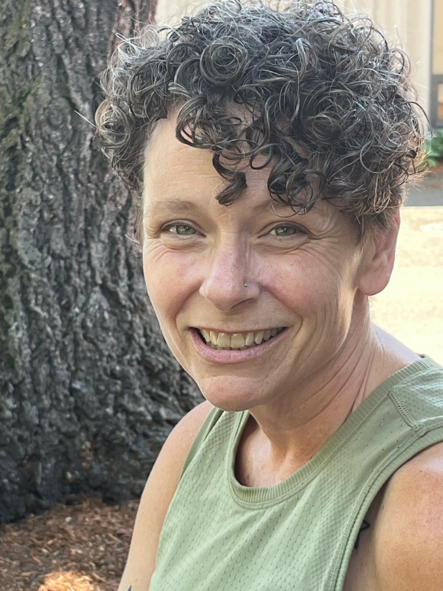 Woman with short curly hair smiling outdoors near a tree.