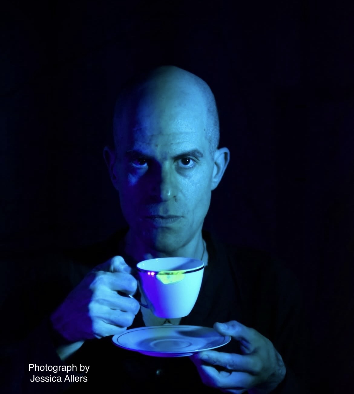 Person with shaved head holding a cup and saucer, illuminated by blue light, dark background.