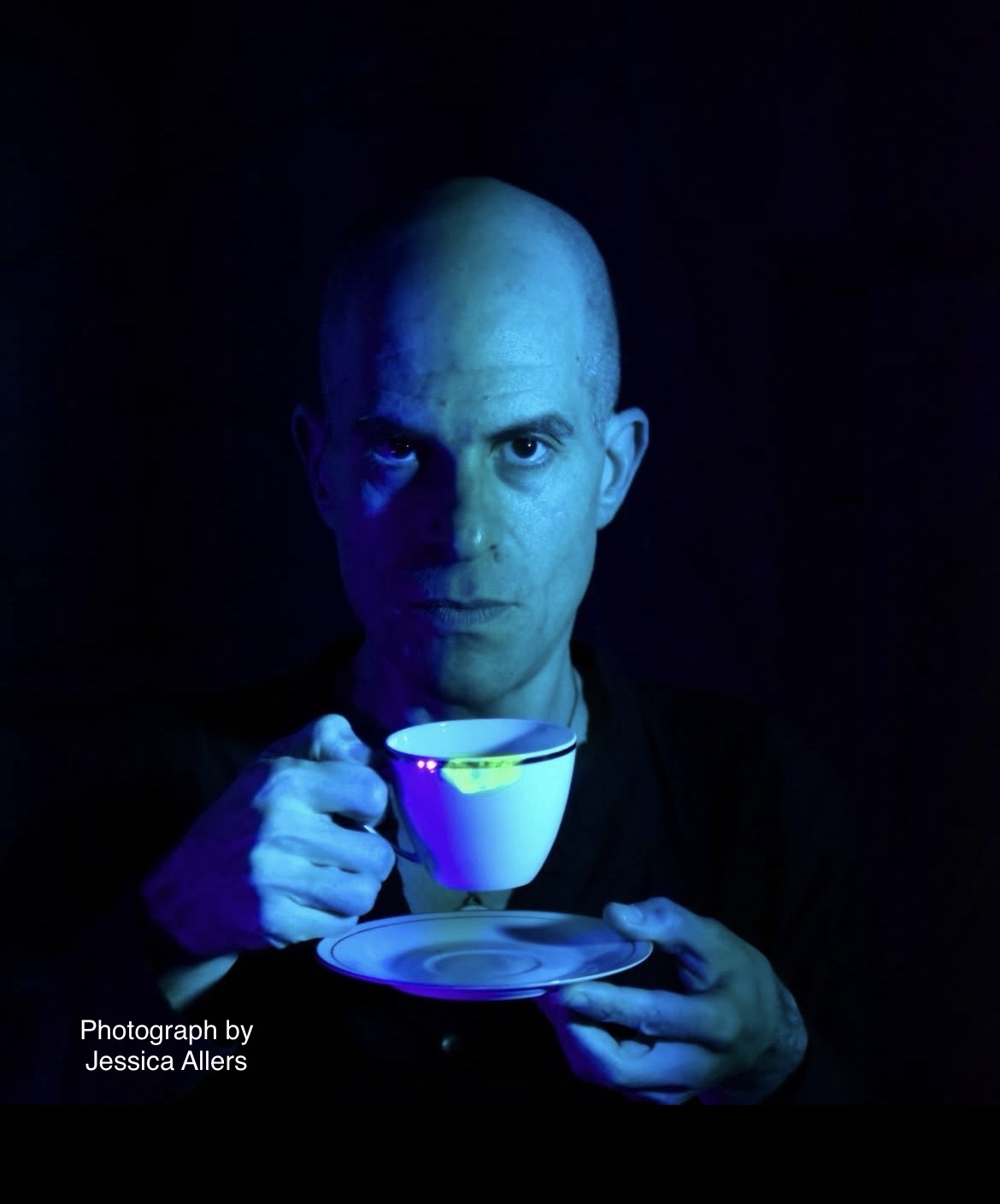 Bald person holding a teacup and saucer, illuminated with blue light, against a dark background.