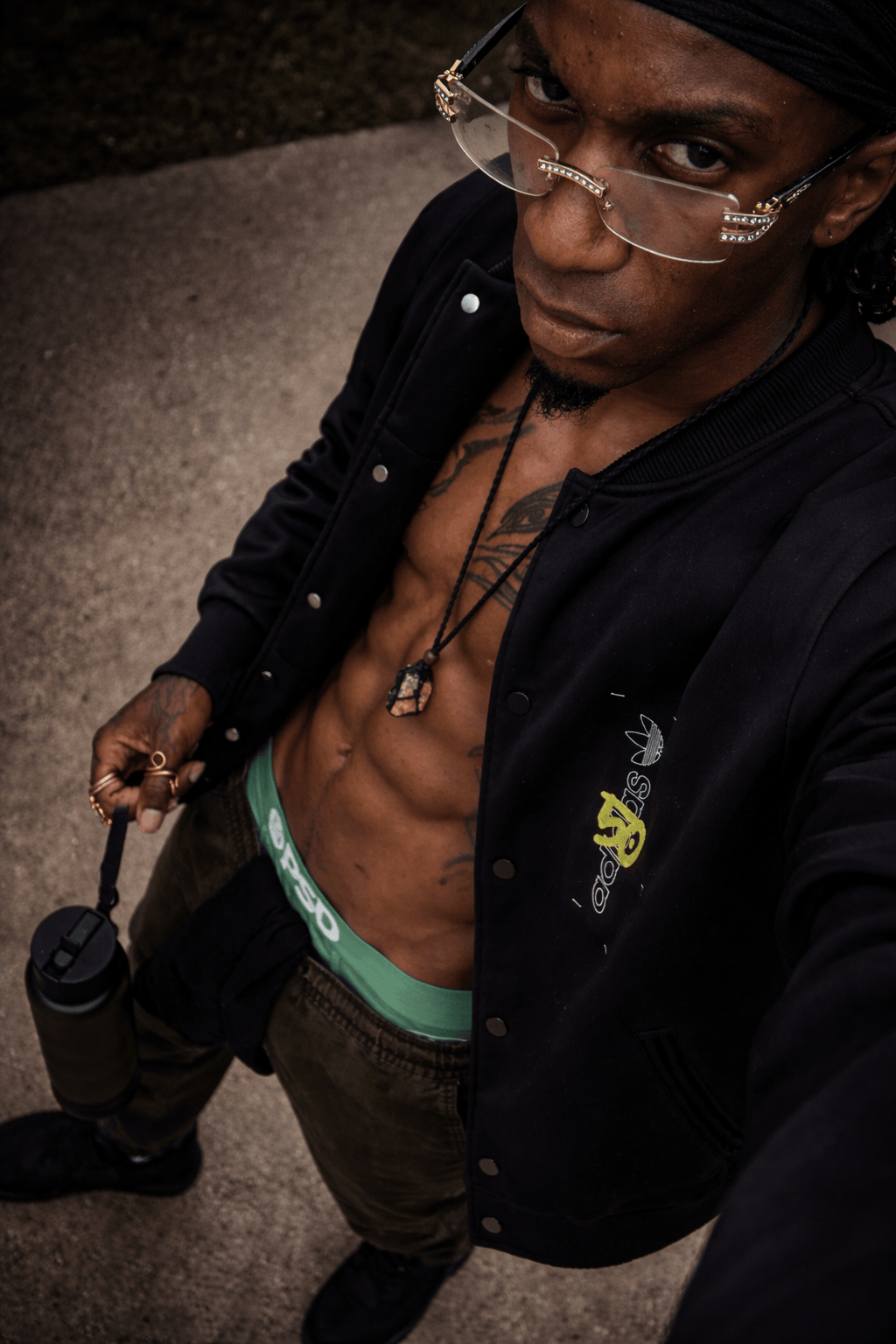 Person with glasses, black head covering, open black jacket, showing chest and abs, holding a camera, standing outdoors.