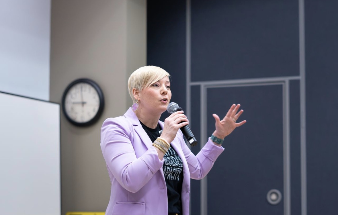 Woman with short blonde hair speaking into a microphone in a room with a clock on the wall.