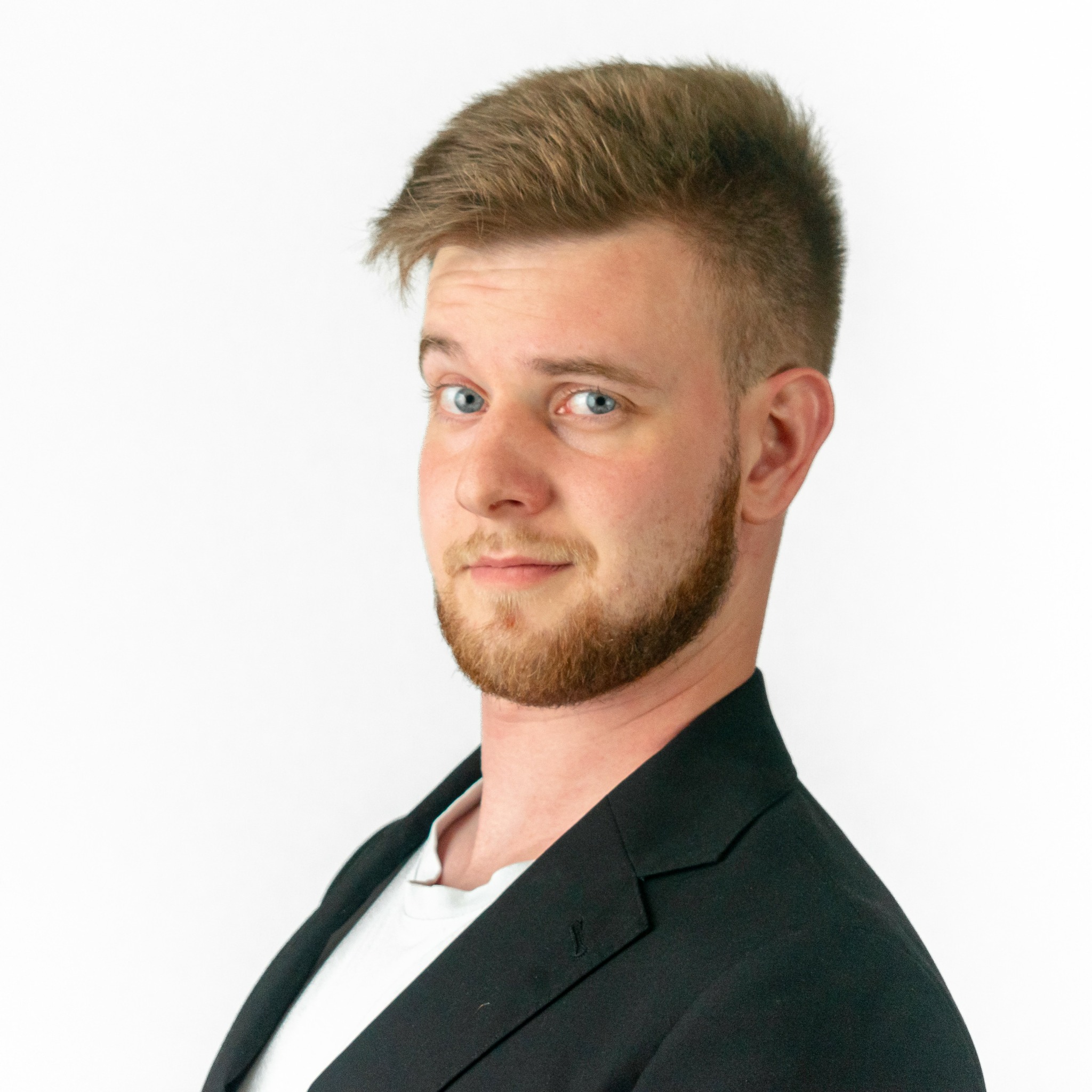 Young man with short, light brown hair and a beard, wearing a black blazer and white shirt, looking sideways.
