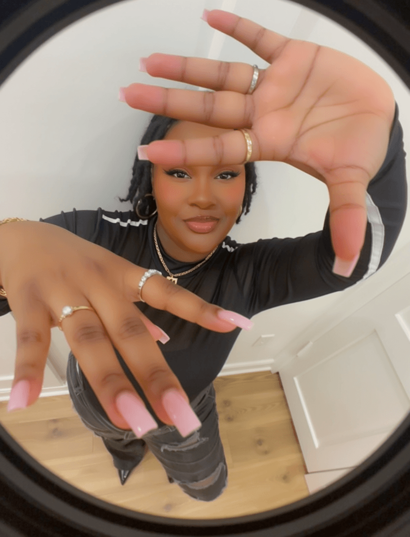 Woman with long pink nails and rings, holding hands up in front of camera, standing indoors.