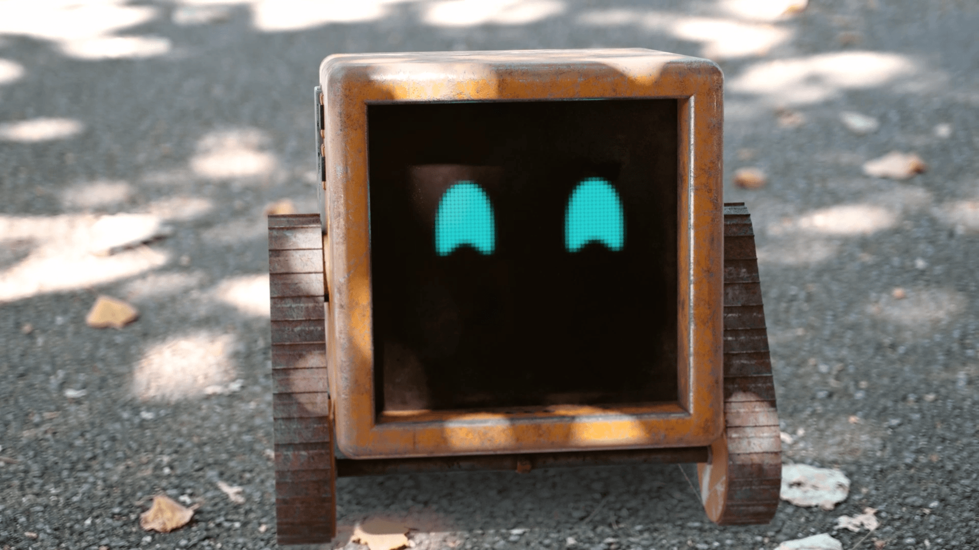 A small wooden robot with a black screen displaying two blue eyes, on wheels outdoors with fallen leaves.