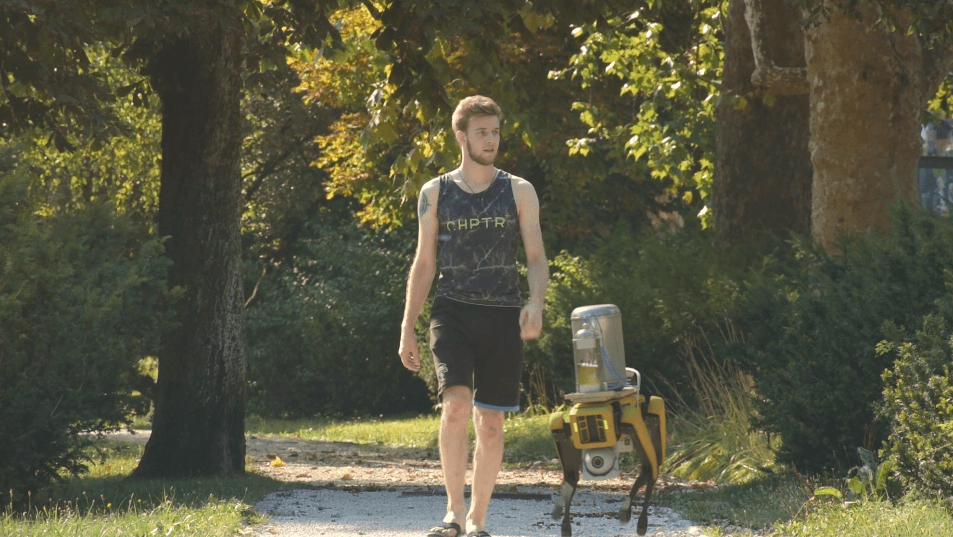 Young man walking on a path in a park with a yellow robot nearby, trees and sunlight in the background.