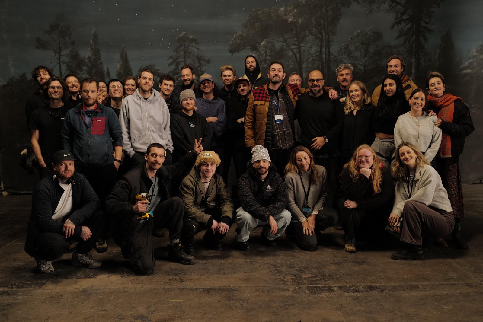 Group of people posing indoors, some standing and some kneeling, with a painted backdrop of trees and sky.