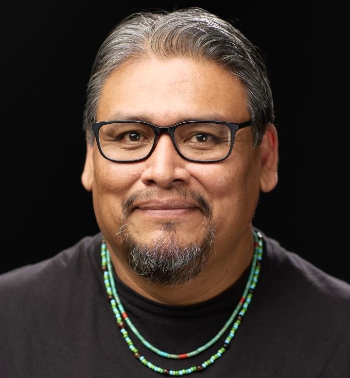 Man with glasses, gray hair, beard, wearing black shirt and beaded necklaces, smiling against black background.