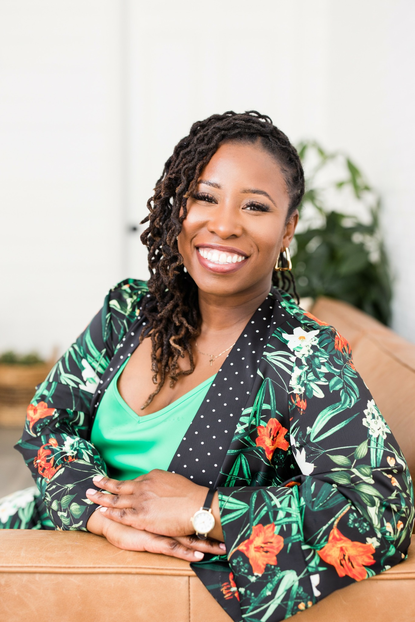 Smiling woman with curly hair, wearing a floral jacket and green top, sitting at a table with a plant in background.