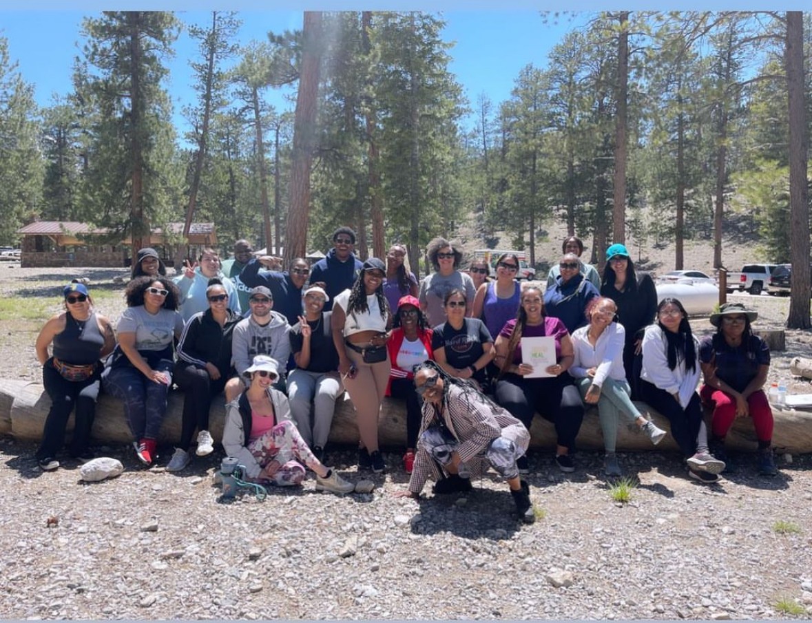 Group of people outdoors in a forested area, some sitting and some standing, with trees and parked cars in background.