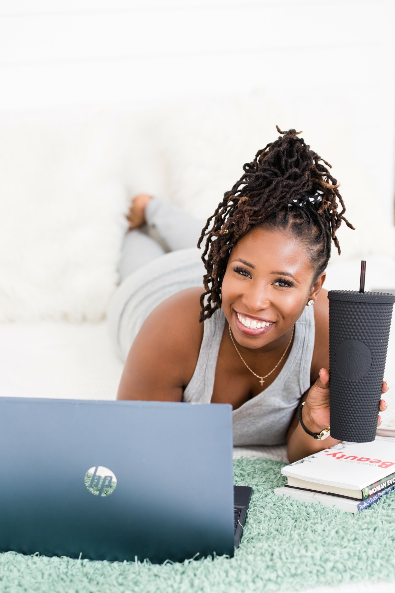 Smiling woman with dreadlocks lying on her stomach with a laptop, holding a large drink, on a green rug.