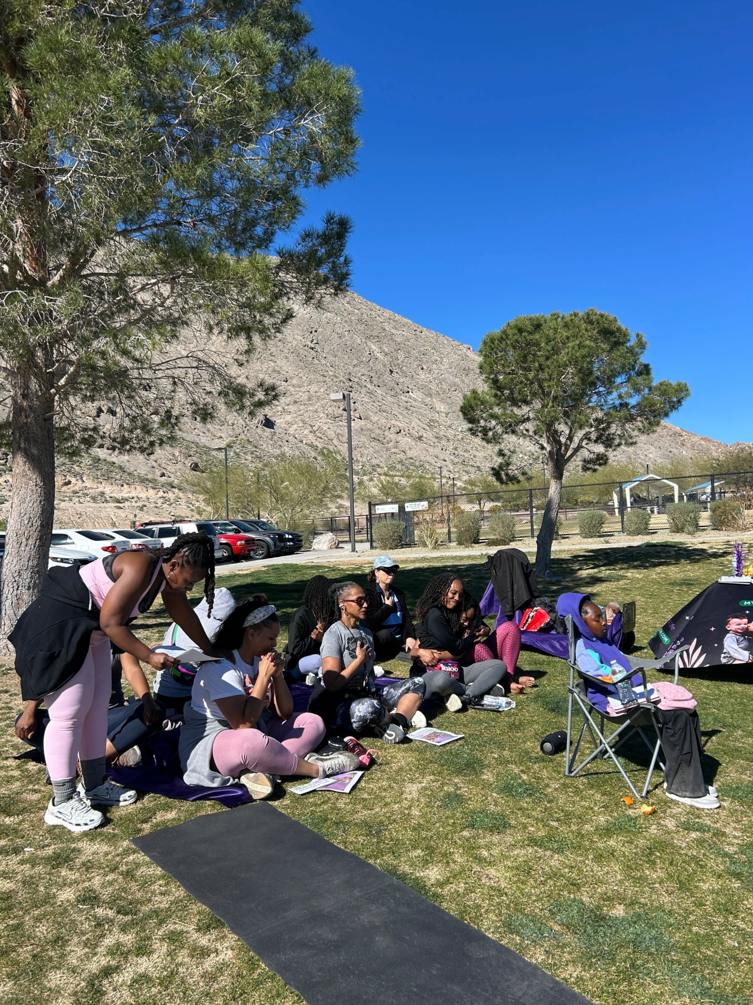 Group of people sitting on the grass with backpacks and chairs outdoors, trees and a hill in the background.