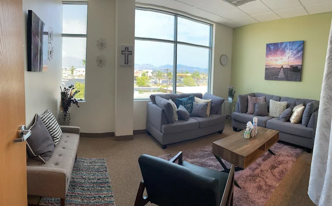 Living room with large windows, sofas, a coffee table, and a chair, decorated with artwork and plants.