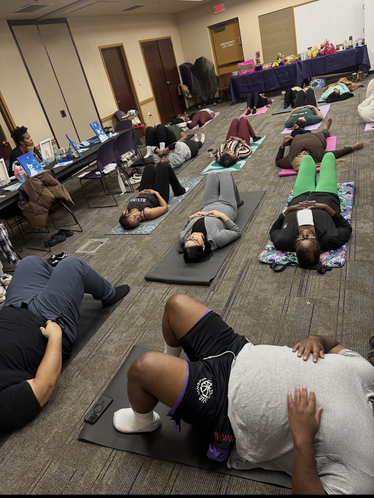 People lying on mats in a room, participating in a group exercise or yoga session, with tables and chairs in the background.