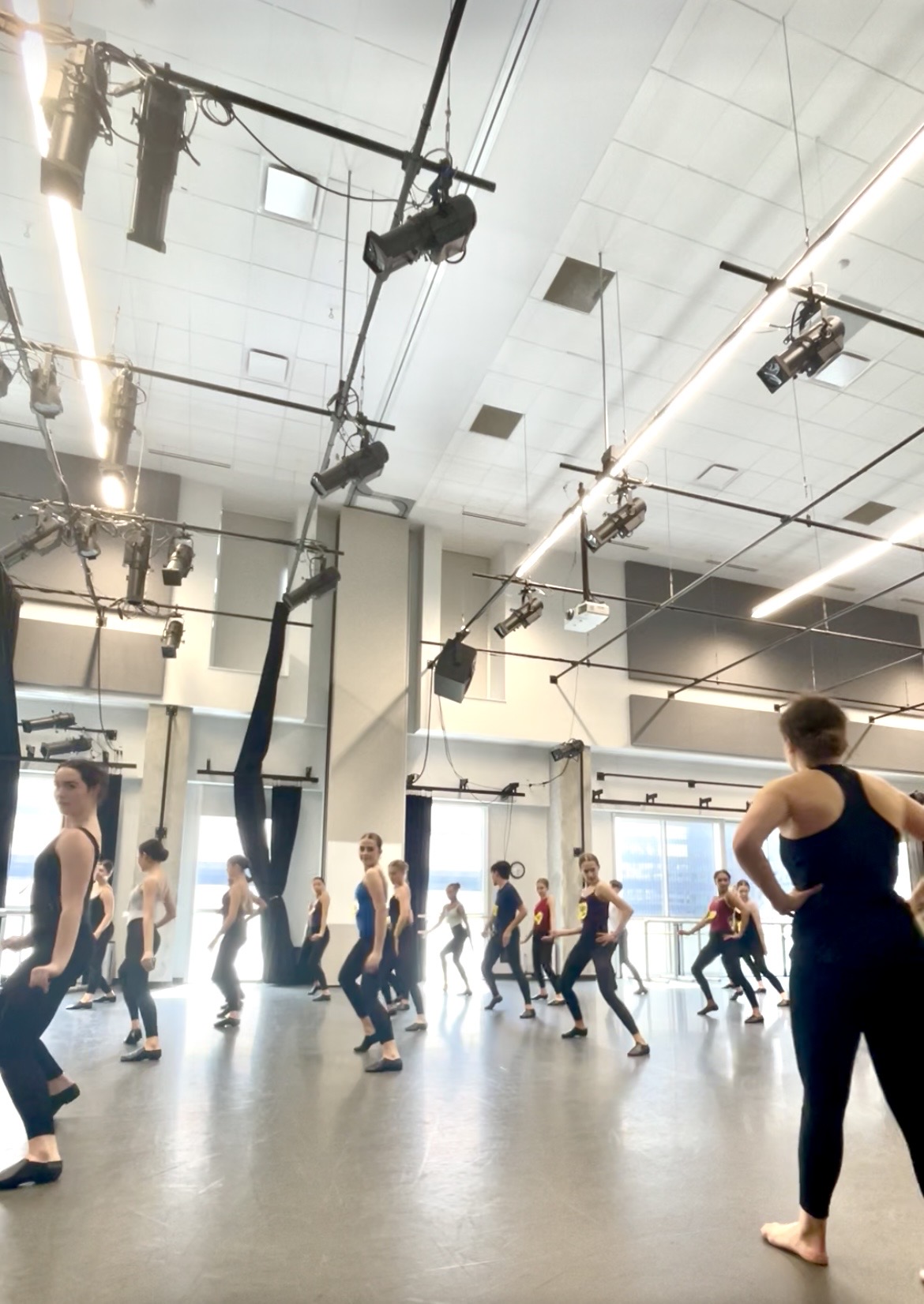Group of people practicing aerial silk dance in a spacious studio with high ceiling and lighting fixtures.
