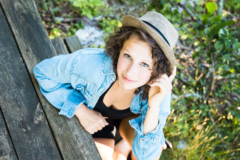 Young woman with curly hair wearing a hat and denim jacket, sitting outdoors among greenery, smiling at the camera.