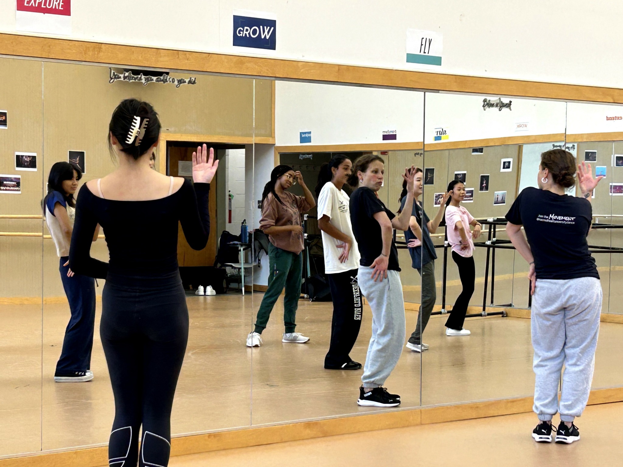 Group of women practicing dance or exercise in a studio with a large mirror, some raising hands, others observing.