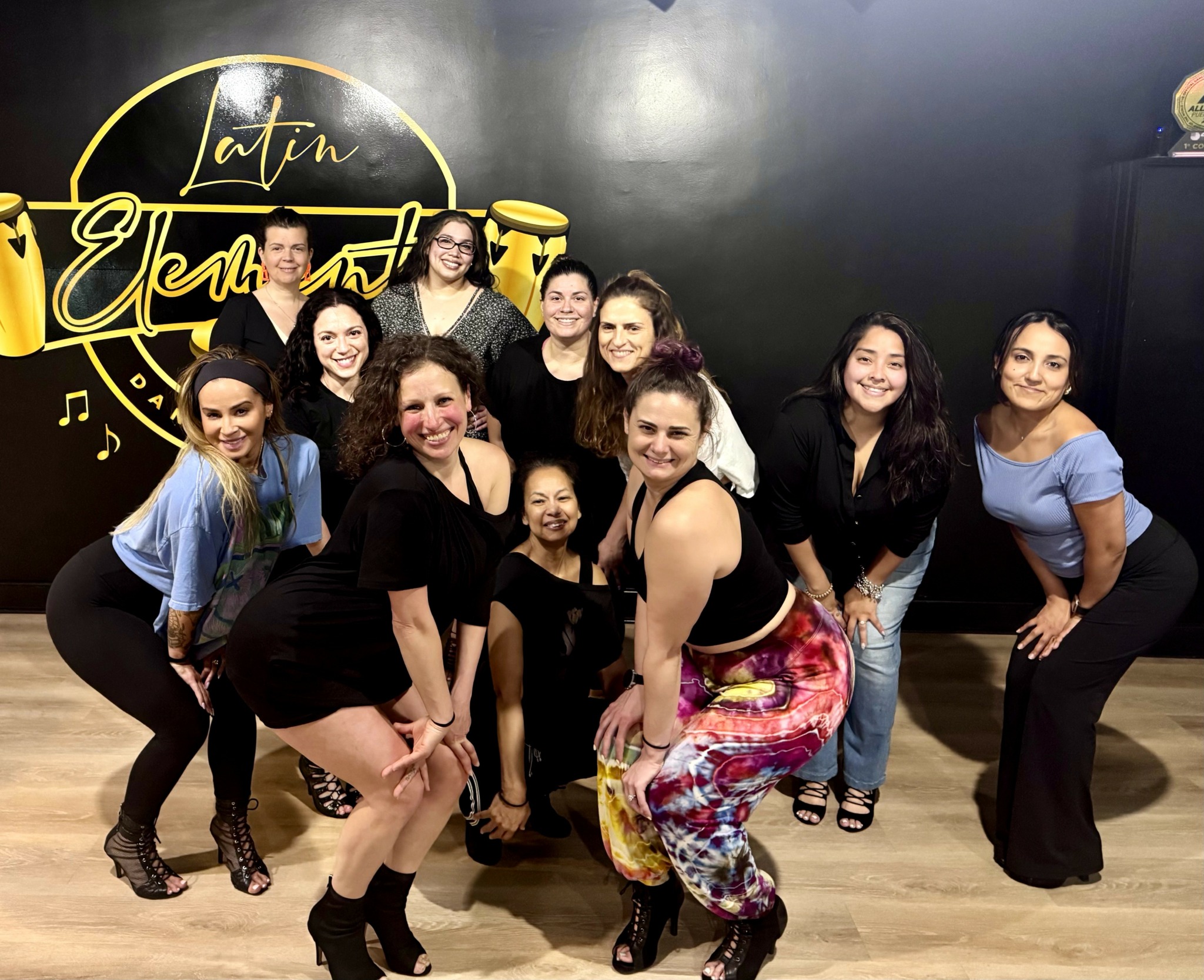 Group of ten women posing together in a dance studio with a black wall and gold logo background.