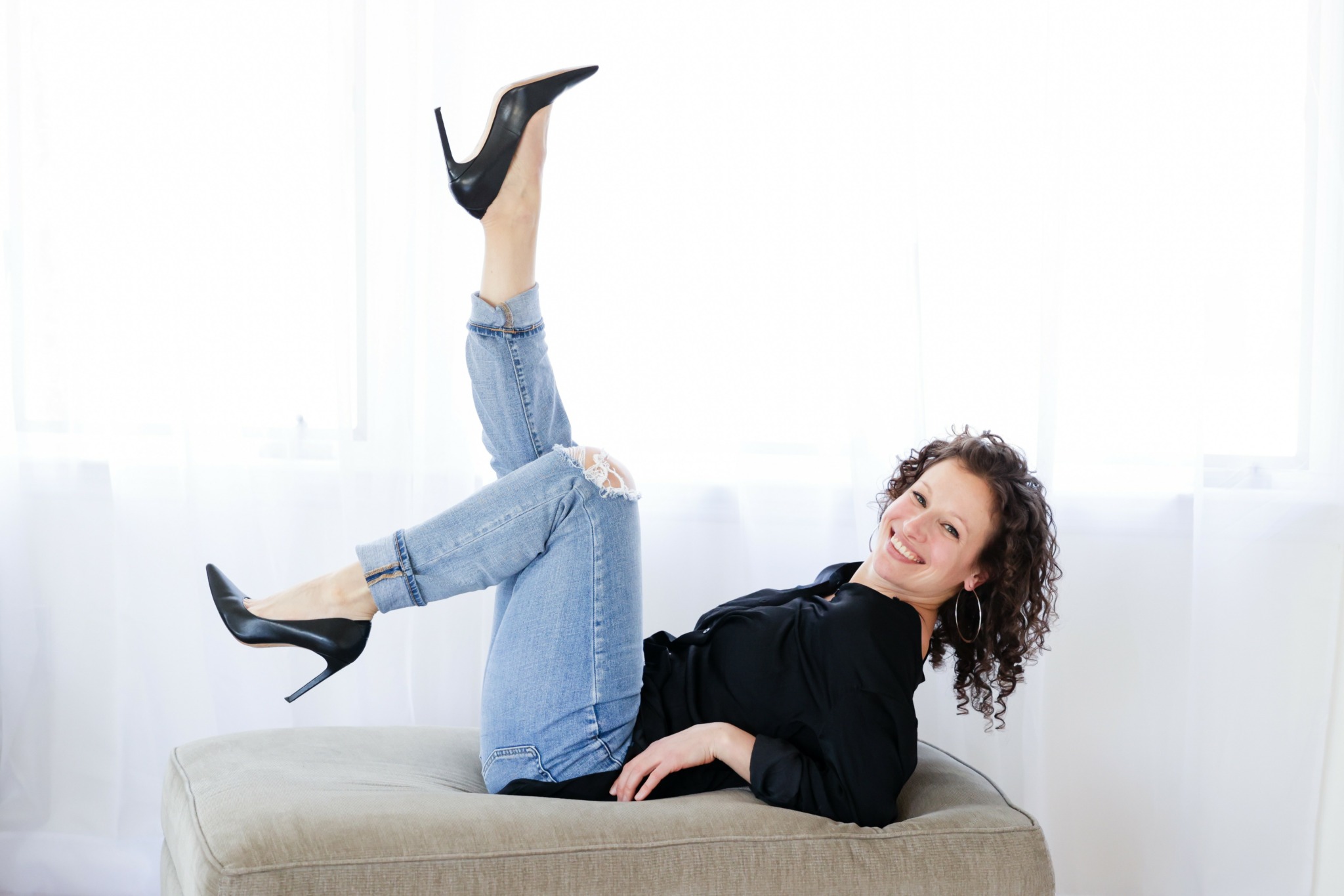 Woman lying on a bench with legs raised, smiling, in a bright room with white curtains.