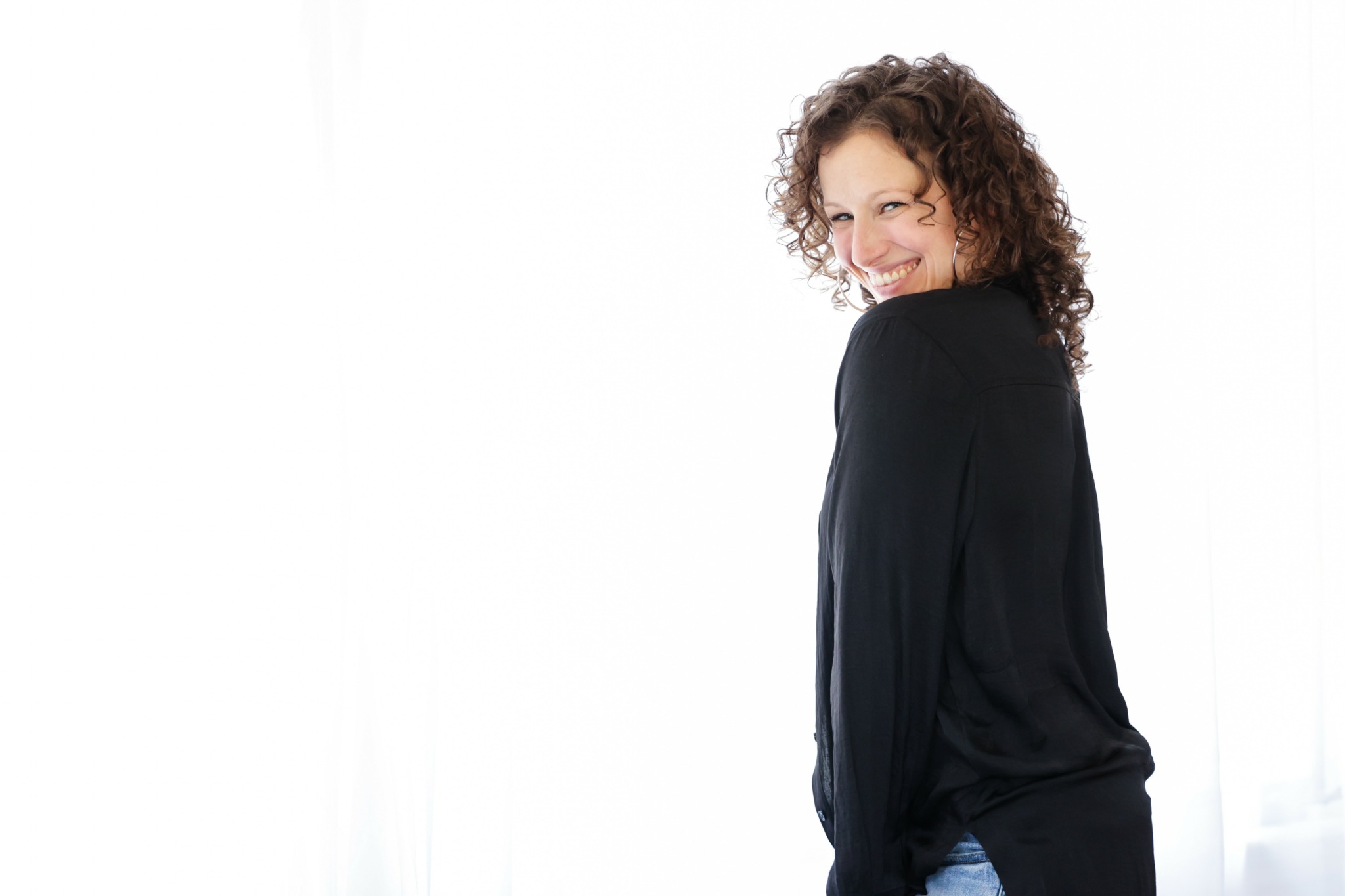Woman with curly hair smiling, wearing a black top, standing in front of a white background.