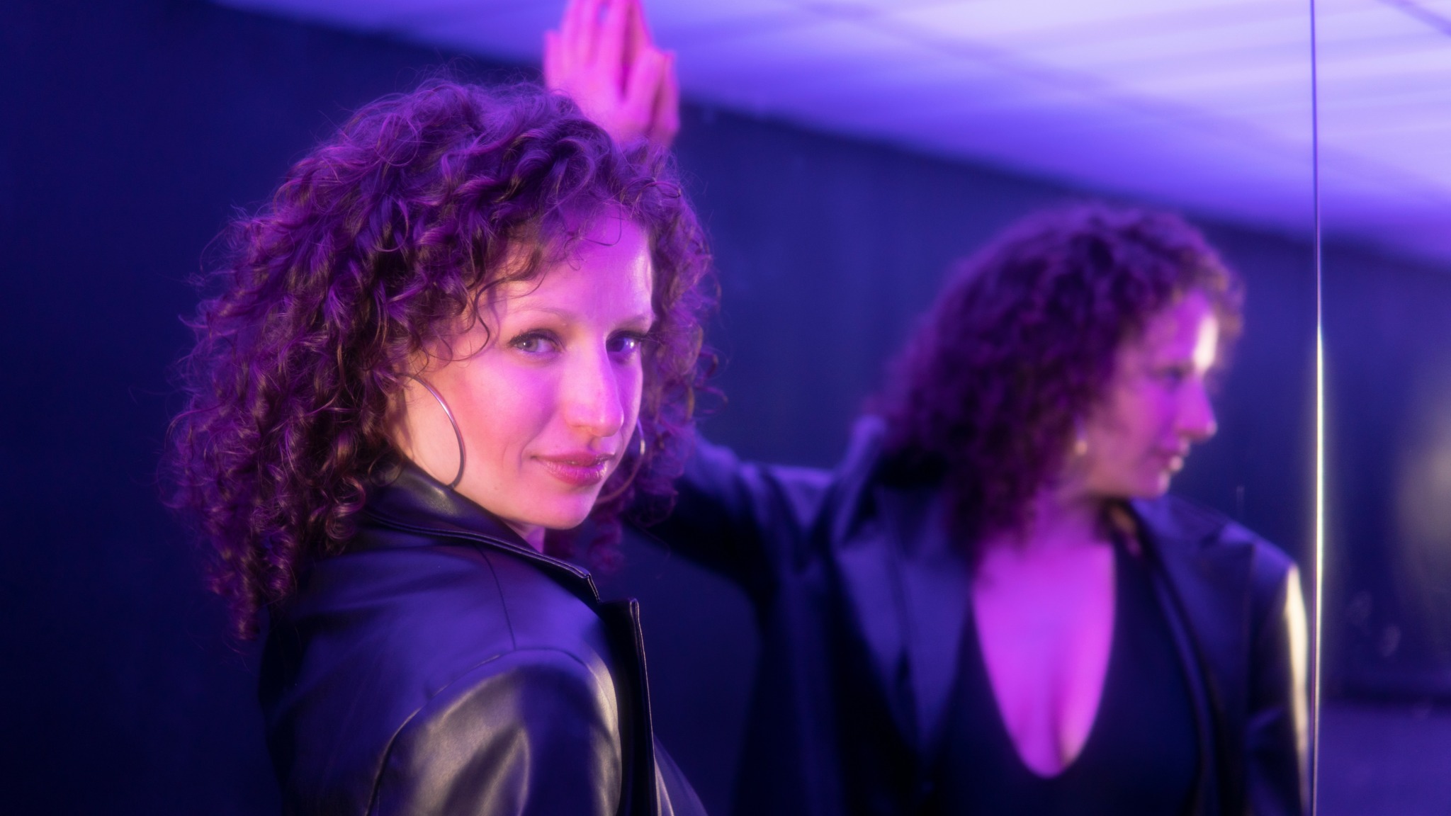 Woman with curly hair looking at mirror, purple lighting, wearing black jacket, in a room with a mirror.