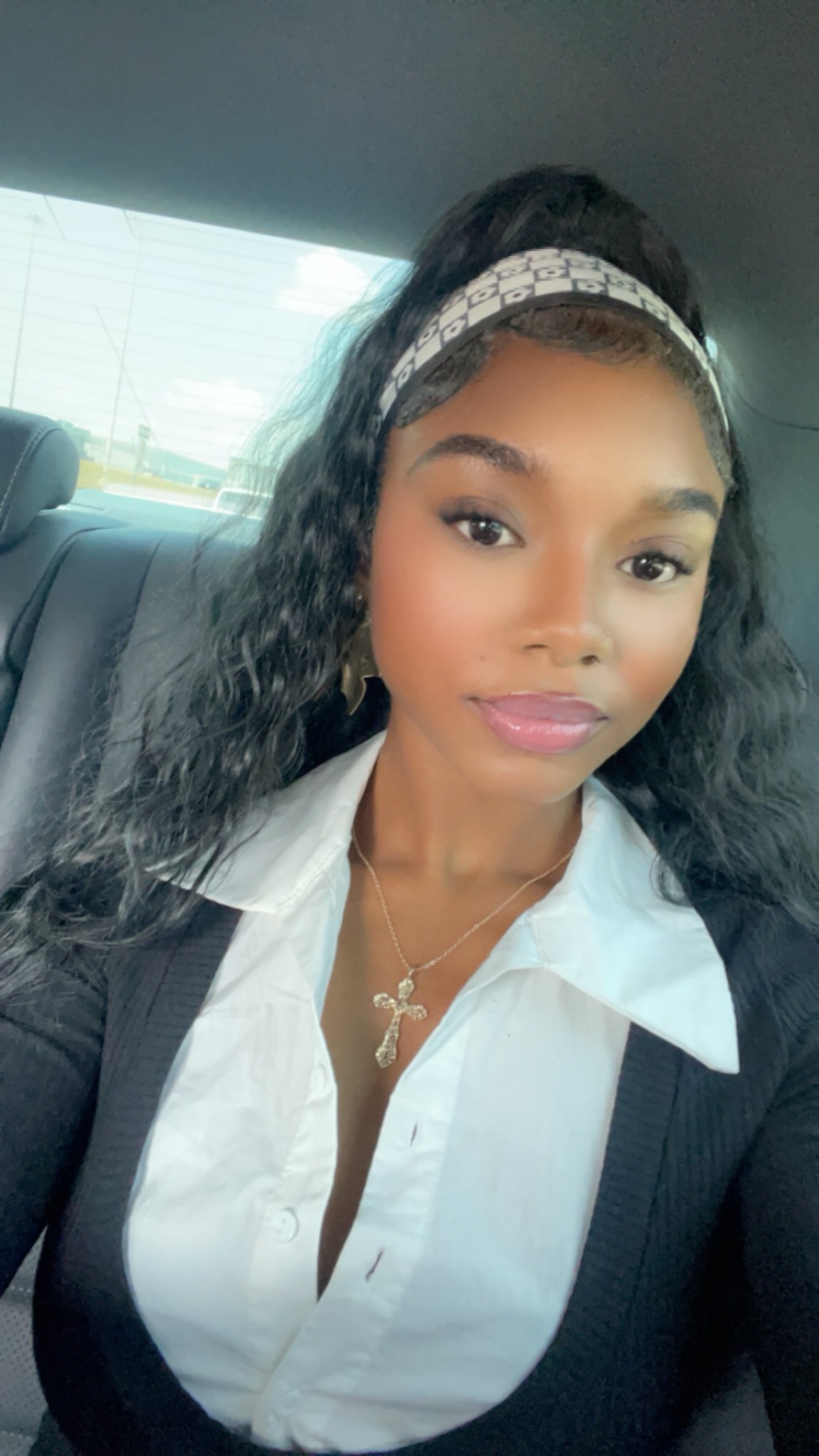 Young woman with curly hair wearing a white shirt, black blazer, and a headband, sitting in a car.
