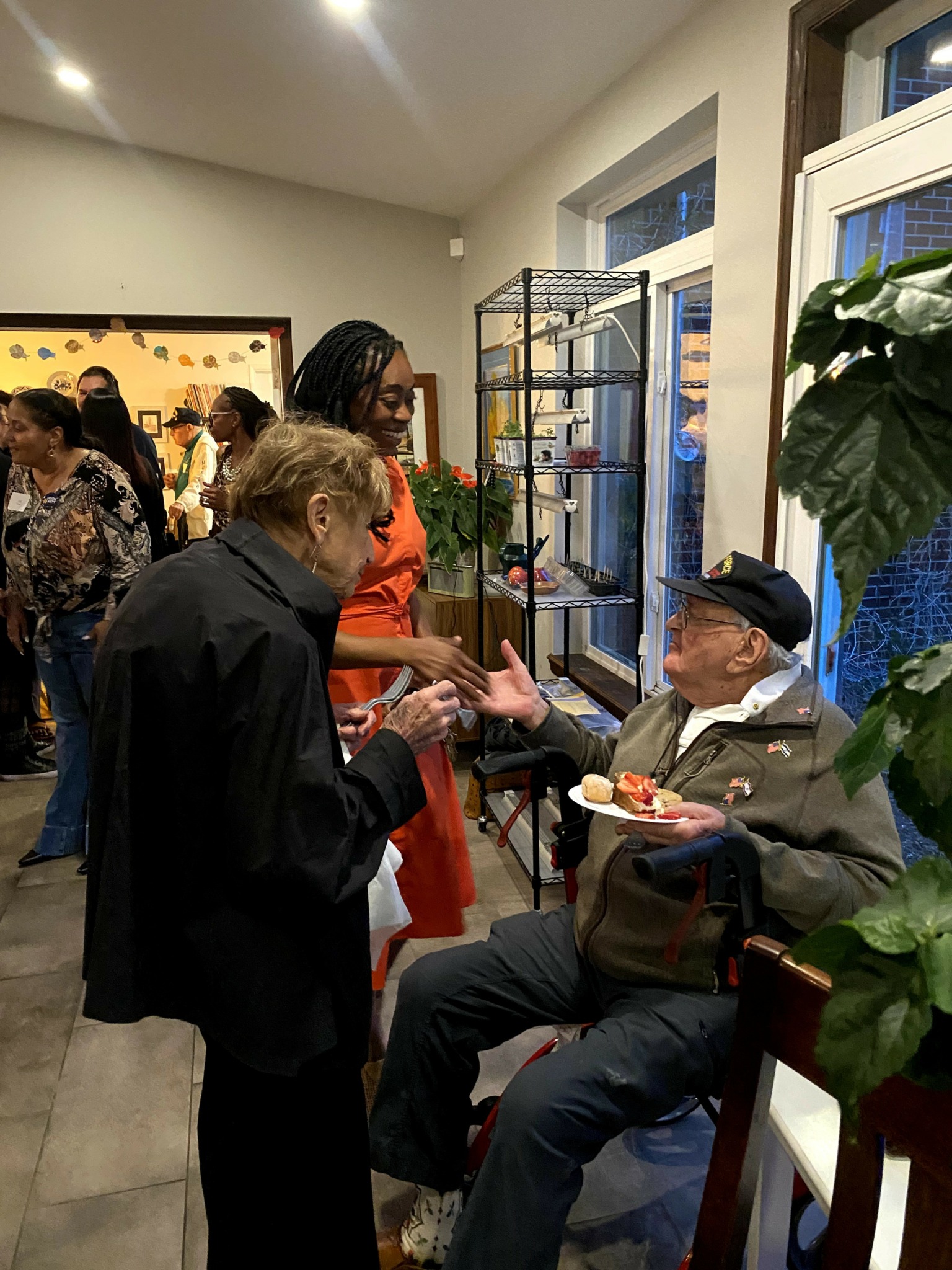 Three people engaged in conversation indoors, one seated holding a plate, two standing, with a window and plants nearby.