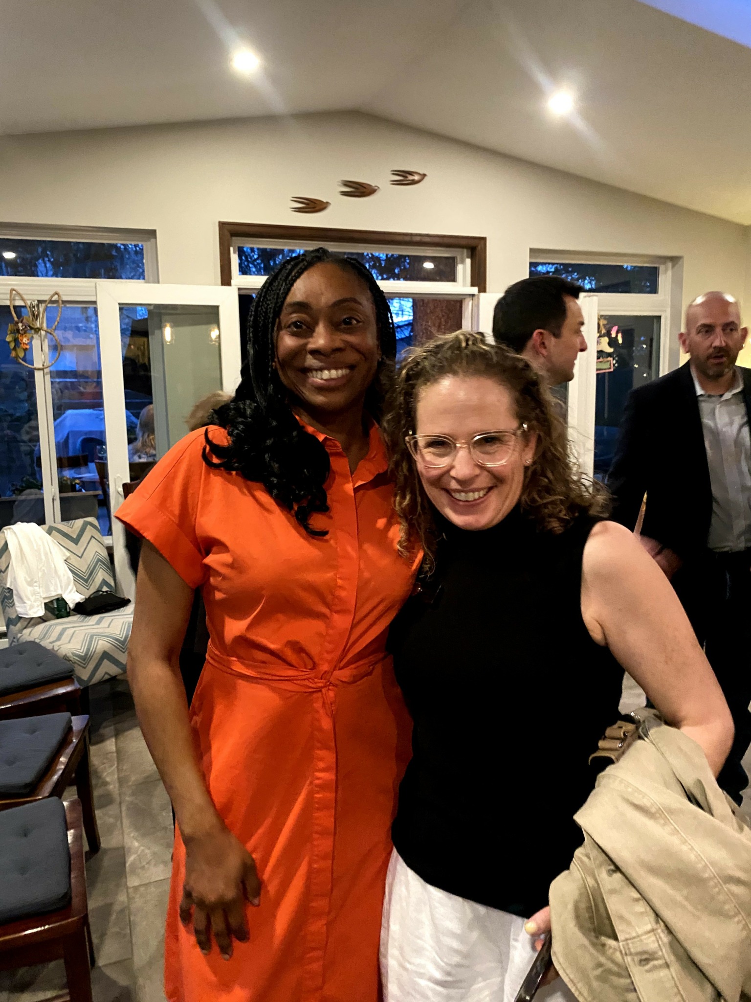 Two women smiling indoors, one in an orange dress and the other in a black top and white pants.