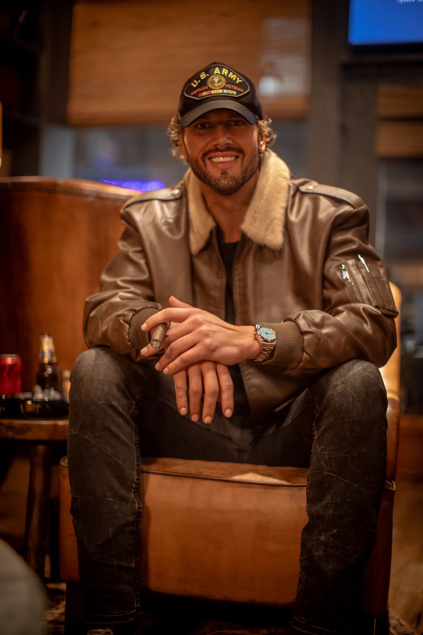Man with a beard and glasses sitting on a chair, wearing a leather jacket and cap, smiling.