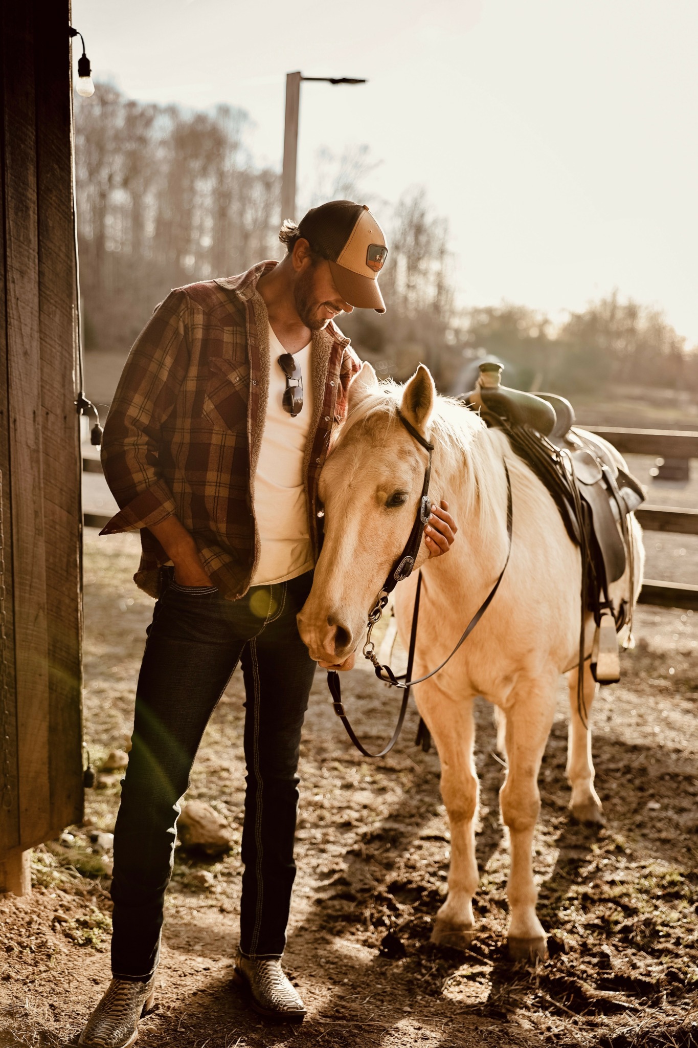 Person in plaid shirt and cap standing next to a white horse with saddle outdoors during daytime.