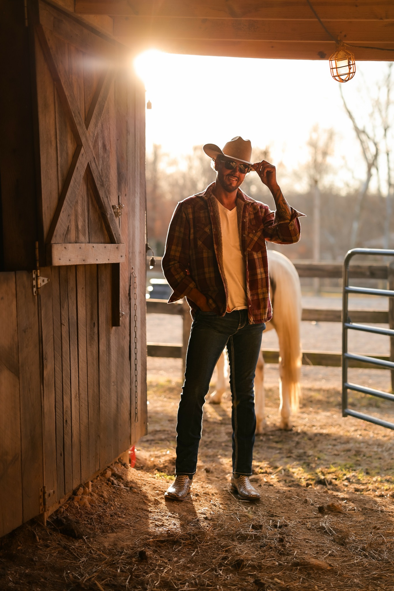 Young man wearing a cowboy hat, plaid shirt, and jeans standing near a horse in a barnyard during sunset.