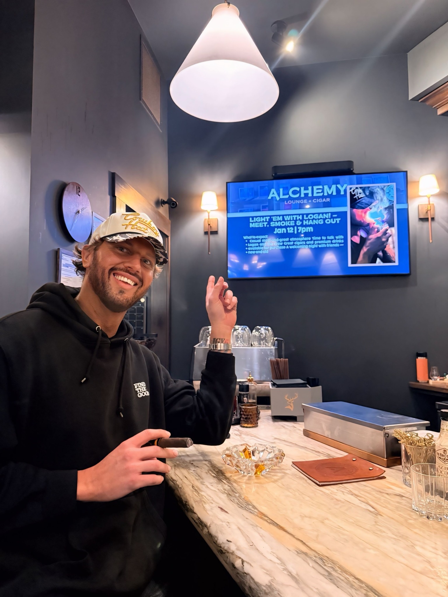 Smiling person wearing glasses and a cap, sitting at a bar with a marble countertop, pointing upward.