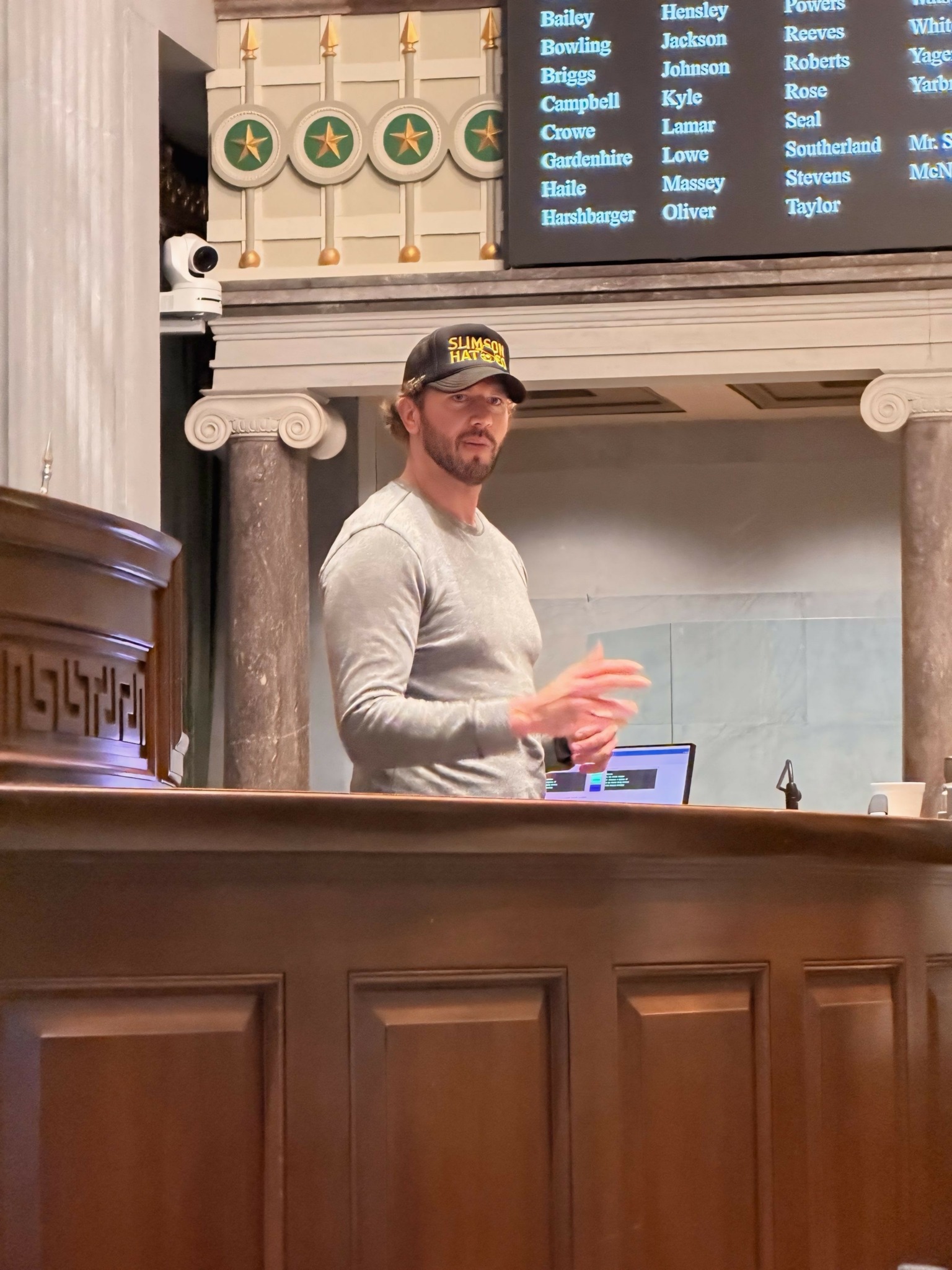 Man with beard and glasses wearing a cap and gray shirt standing behind a wooden counter inside a building.