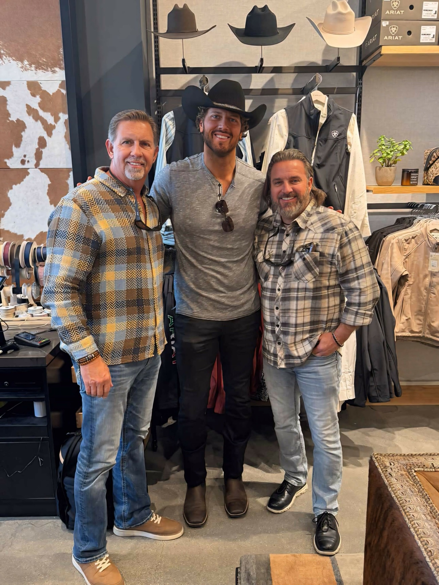 Three men stand together inside a clothing store, smiling, with hats displayed on shelves behind them.