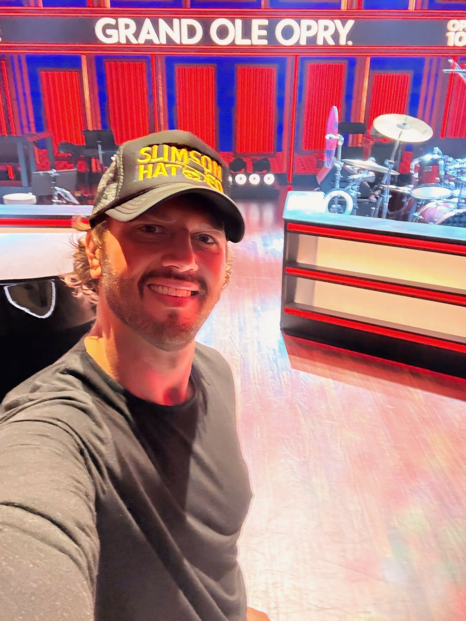 Man taking a selfie in a theater with red and blue decor and a sign reading 'Grand Ole Opry'.